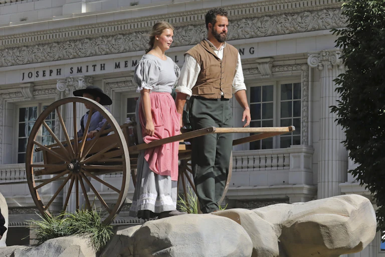 A young woman and man with a beard in old-fashioned pioneer clothing pose with a wooden handcart.