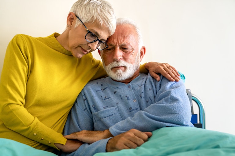 A woman with short gray hair and glasses wearing a yellow sweater with her arm around a man with gray hair and bear.