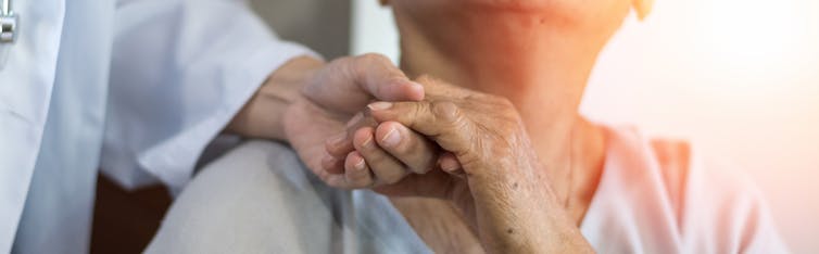 Cropped image of a person in a white coat standing behind a seated older person, holding their hand