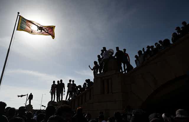 Protesters are seen on top of presidential secretariat whil a flag flies in the foreground. The sun is setting behind them.