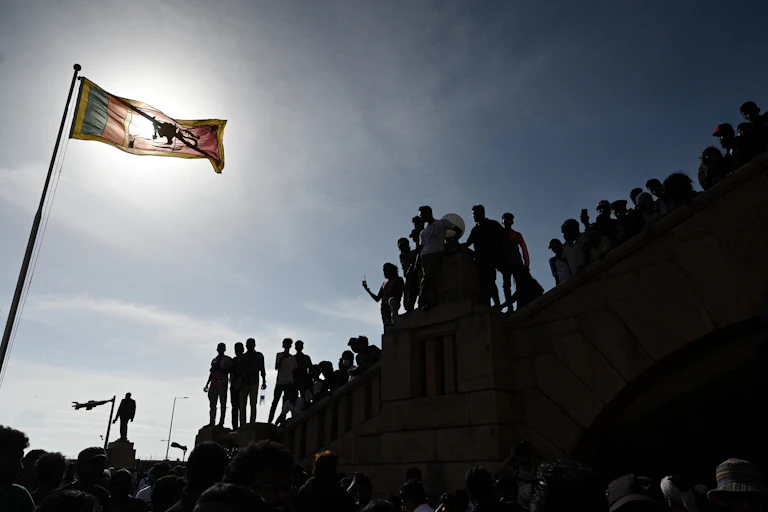 Protesters are seen on top of presidential secretariat whil a flag flies in the foreground. The sun is setting behind them.