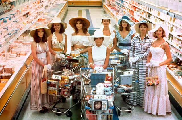 smiling women in dresses, hats and gloves with trolleys in a supermarket aisle
