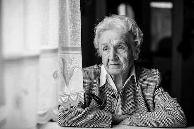Foto en blanco y negro de una señora mayor con los brazos cruzados mirando a través de unos visillos.