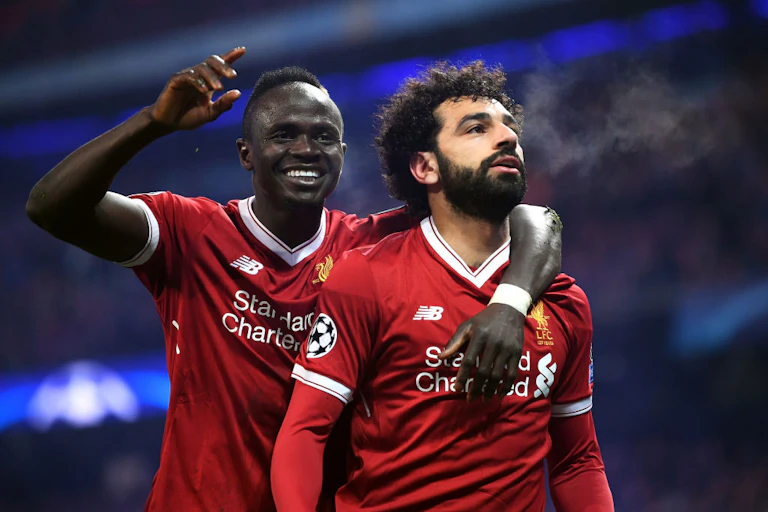 Two men, one with a short mohawk and the other with curly hair embrace, wearing red football jerseys.