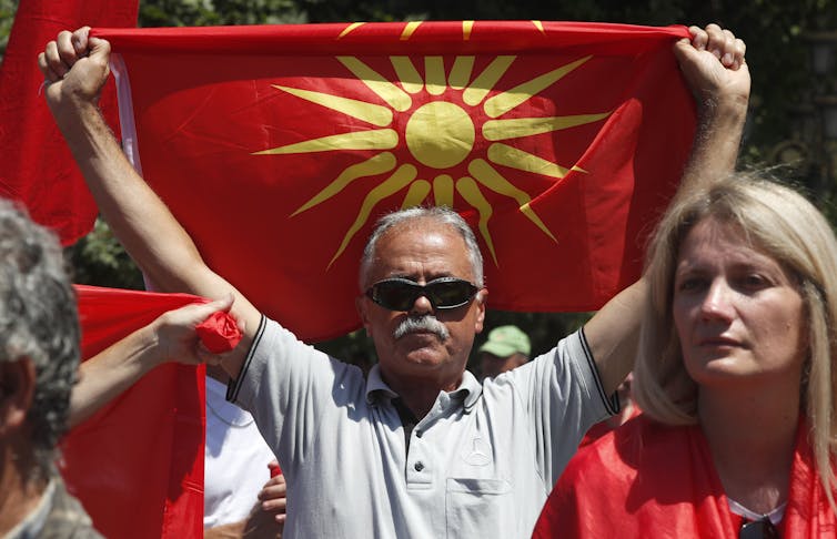 A grey-haired man in sunglasses holds up the Macedonian flag.