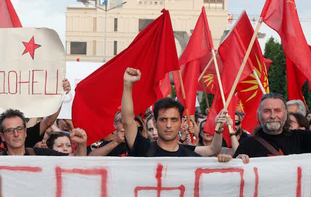 A dark-haired man raises his fist as he marches alongside others waving the Macedonian flag and carrying banners.