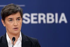 A woman with short dark hair speaks into a microphone. The word Serbia is seen in the background.