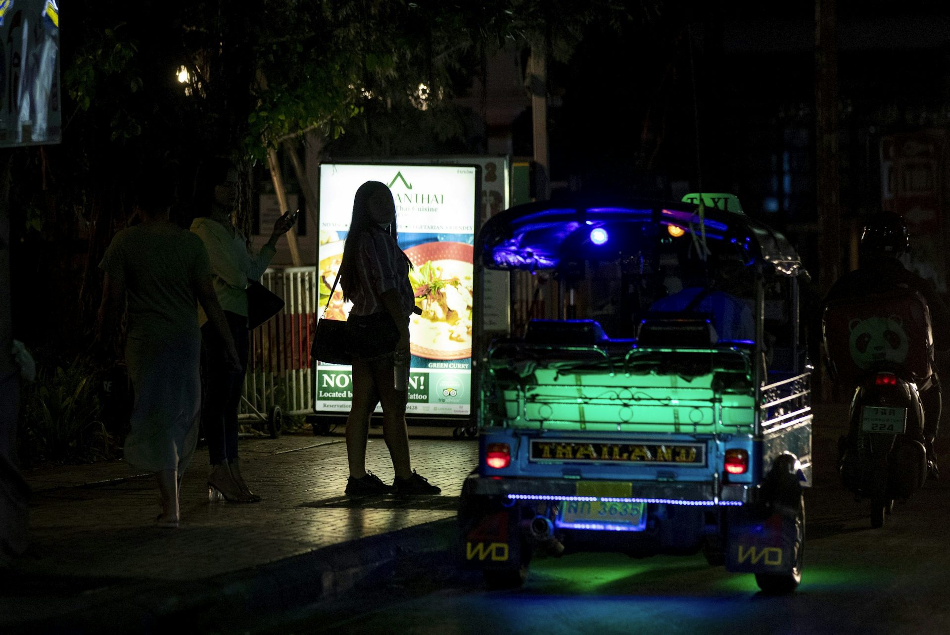 A woman stands backlit next to a dimly lit bus that reads 'Thailand' with green lighting.