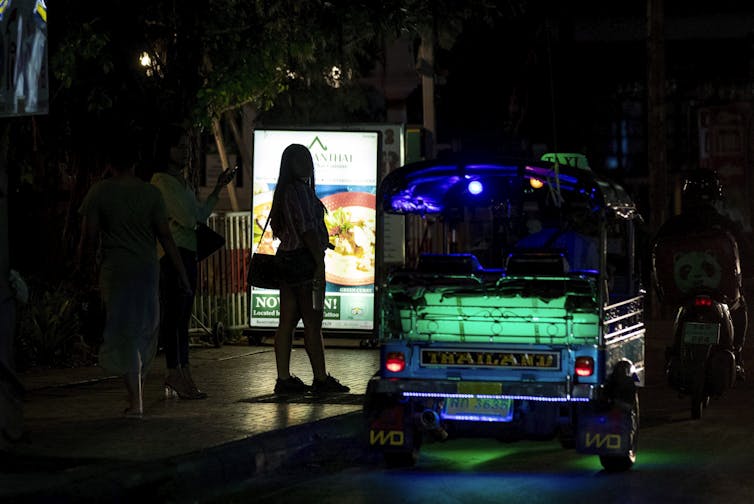 A woman stands backlit next to a dimly lit bus that reads'Thailand' with green lighting.