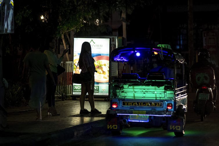 A woman stands backlit next to a dimly lit bus that reads 'Thailand' with green lighting.