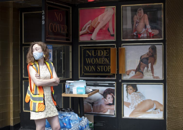 A woman stands wearing a mask with a safety vest on in front of a collage of scantily clad women and a sign that reads'nude women non stop'