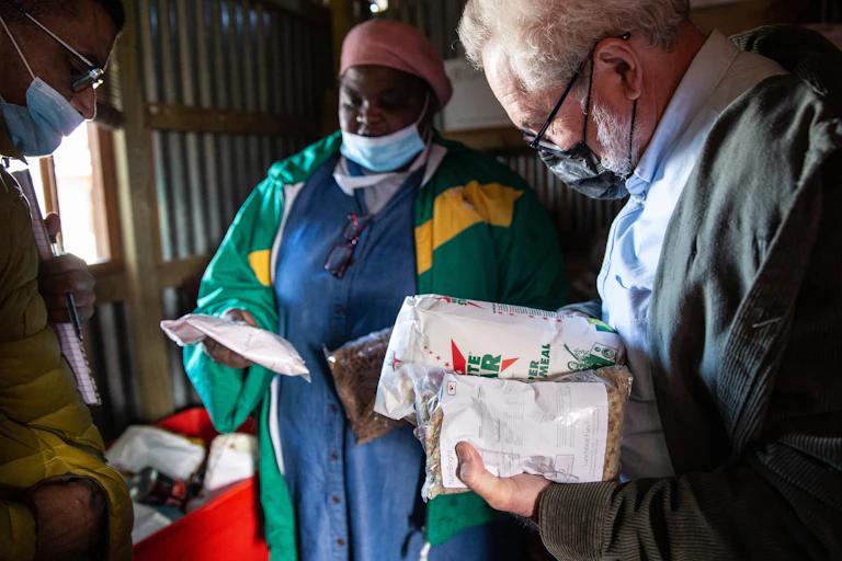 Inside a corrugated iron and wood building, a woman and a man wearing face masks hold packets of maize meal and dry beans
