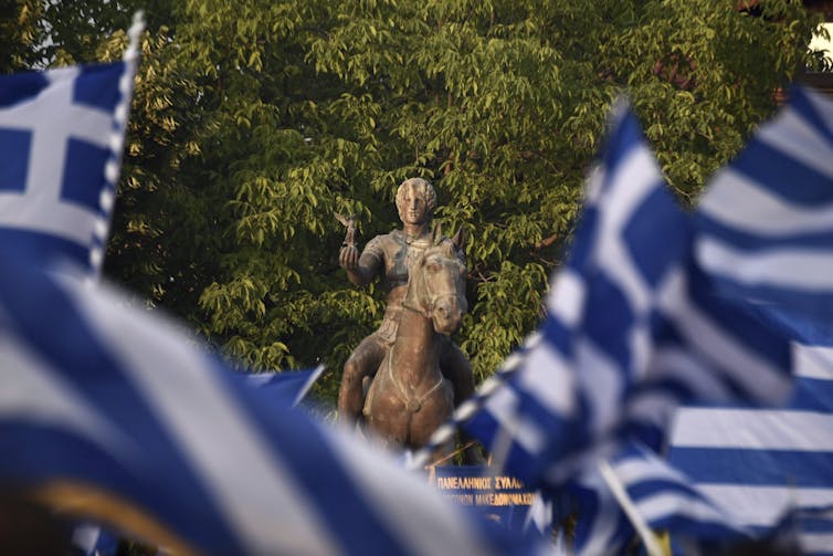 Several blue and white flags frame a statue of Alexander the Great.