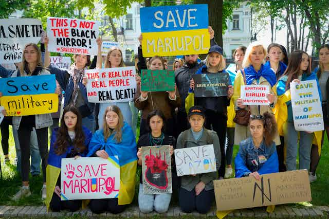 Women hold signs saying Save Mariupol against a backdrop of leafy trees..