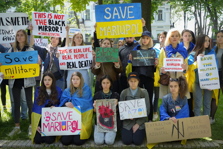 Women hold signs saying Save Mariupol against a backdrop of leafy trees..