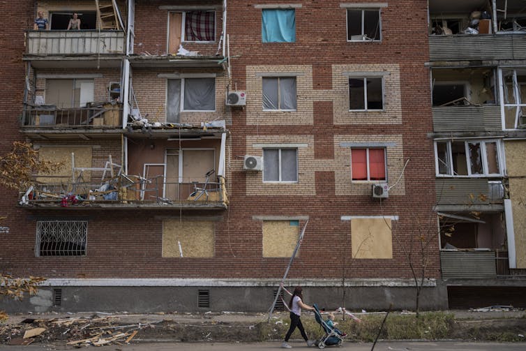 A woman pushes a baby stroller past a damaged apartment building.