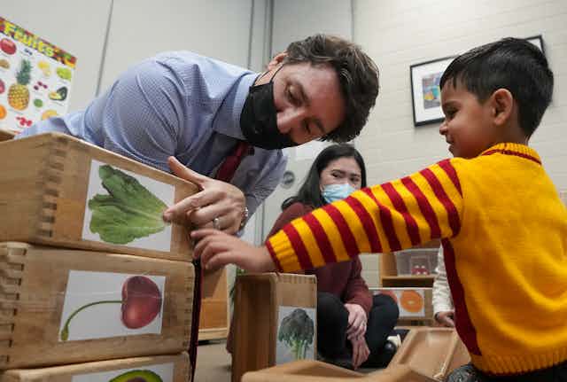 A man in a mask, Justin Trudeau, is seen pointing at a lettuce leaf on a block, while a boy at a child care centre smiles and listens.