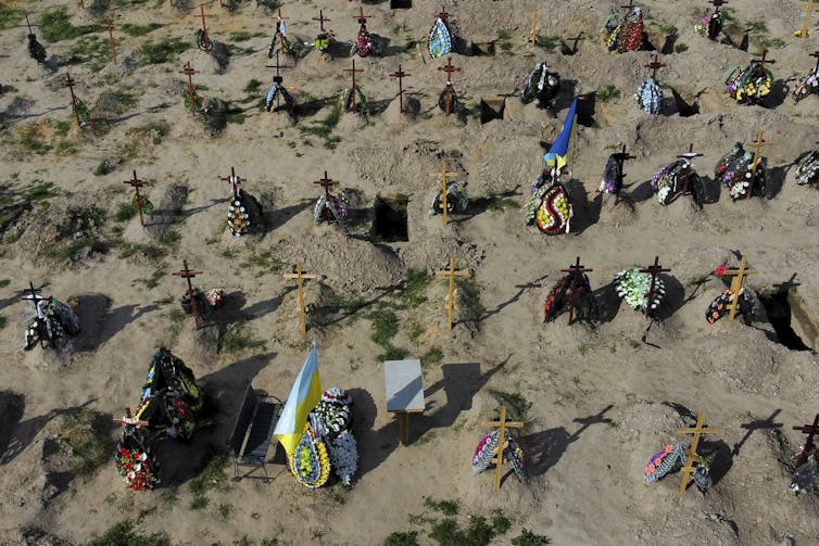 Tombs with crosses are seen in the dirt in a cemetery. Some have flowers, decorations and flags on them.