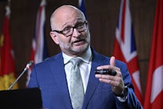 A bald man with a beard and glasses in a navy suit with a light blue tie.