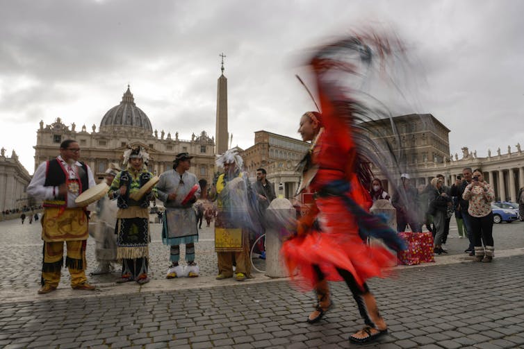 A woman dances in regalia in front of people with handrums