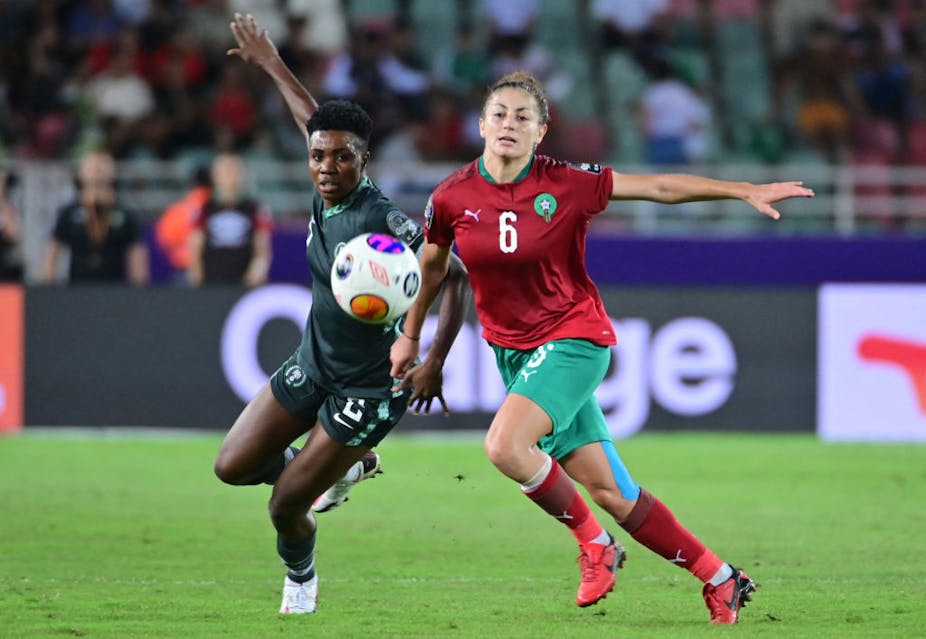 Two women football players tussle, the ball in the air between them, their arms in the air at their sides.