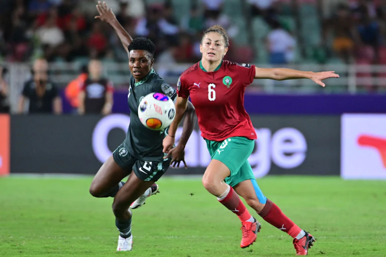 Two women football players tussle, the ball in the air between them, their arms in the air at their sides.