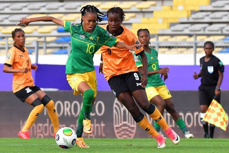 Women footballers battling it out on the field. At the centre two players, one on the ball, her hands at her sides and the other coming from behind.