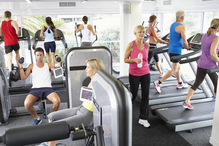 A busy gym full of adults using treadmills, ellipticals and other exercise machines for lifting weights.
