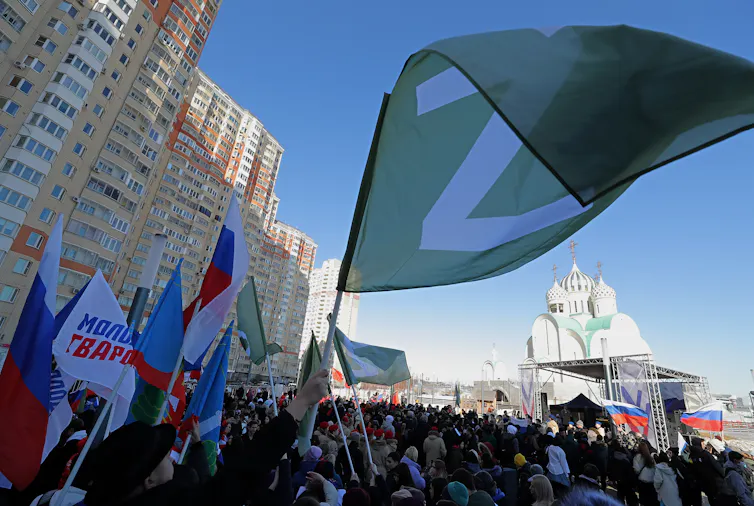 Large crowd outside blocks of flats, one person waving a flag with the letter 'Z'.