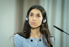 A woman with long dark hair wearing a blue shirt sits with headphones on in front of a microphone.