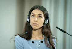 A woman with long dark hair wearing a blue shirt sits with headphones on in front of a microphone.