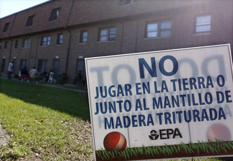 A large brick housing complex with people sitting in lawn chairs outside. A sign on the lawn is in Spanish.
