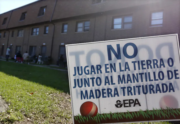A large brick housing complex with people sitting in lawn chairs outside. A sign on the lawn is in Spanish.