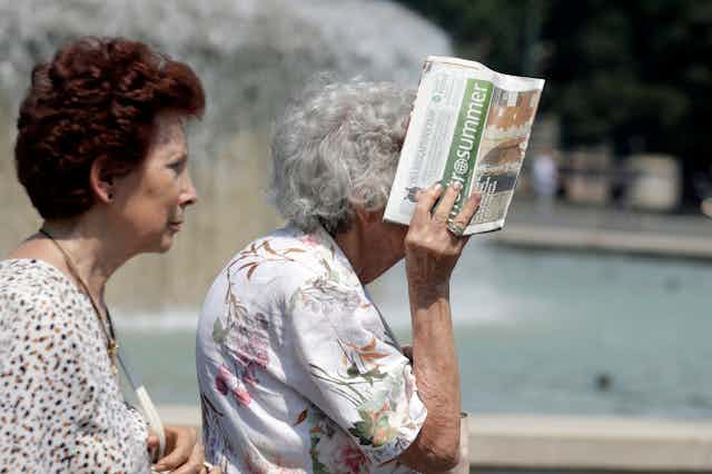 Two older women outside in the sun. One covering her head with a newspaper. 
