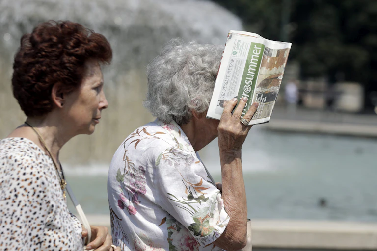 Two older women outside in the sun. One covering her head with a newspaper.
