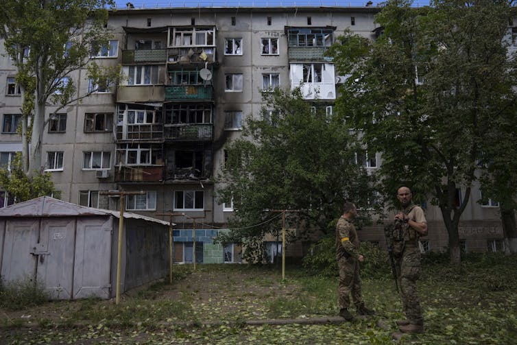Two soldiers stand in front of a charred, 5 story residential building