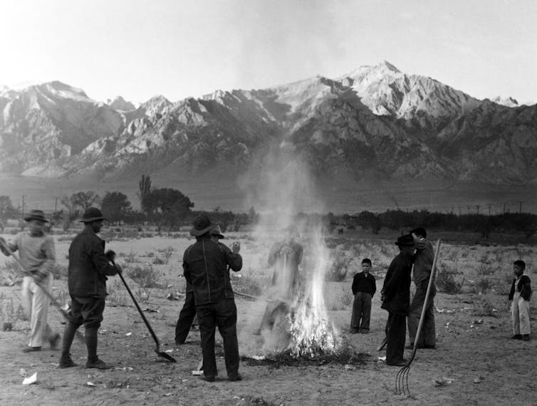 A black and white photo shows people burning leaves in front of a mountain range.