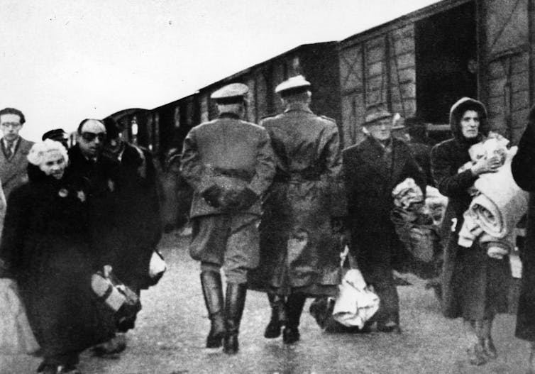 A black and white photo shows the backs of soldiers, facing a small crowd of people carrying suitcases in front of trains