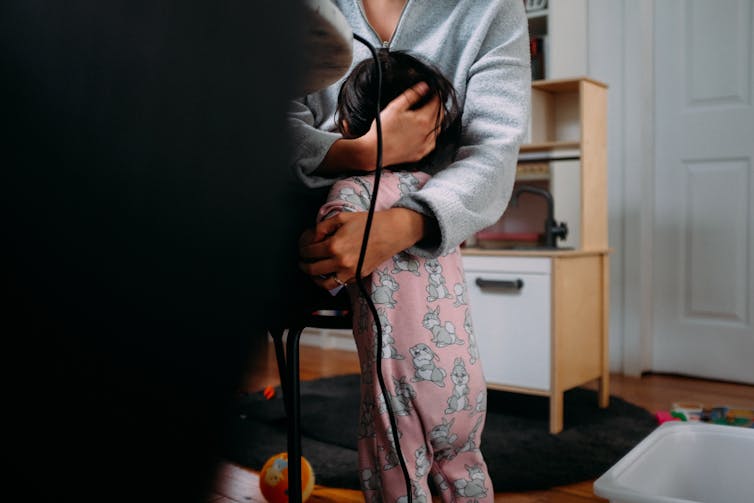 A parent and a baby in a pink and grey sleeper embrace. The adult sits in front of a computer.