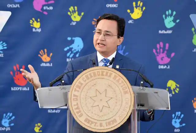 A black haired man with glasses speaks behind a large medallion that says "Seal of the Cherokee Nation" on it.