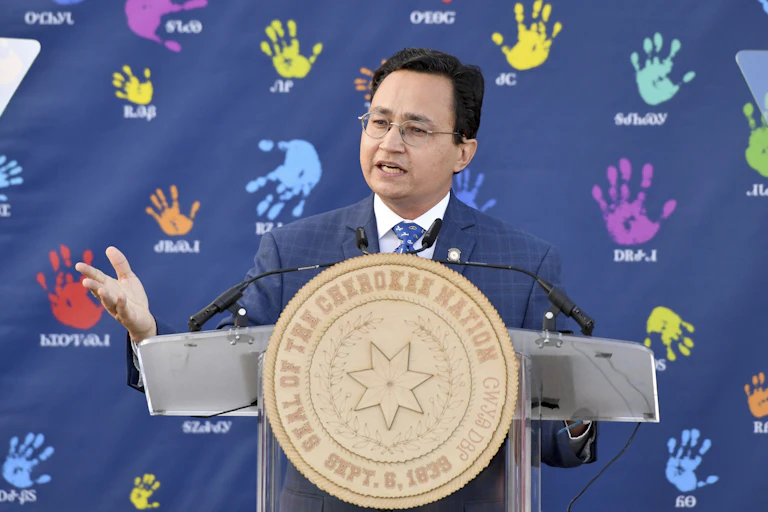 A black haired man with glasses speaks behind a large medallion that says "Seal of the Cherokee Nation" on it.