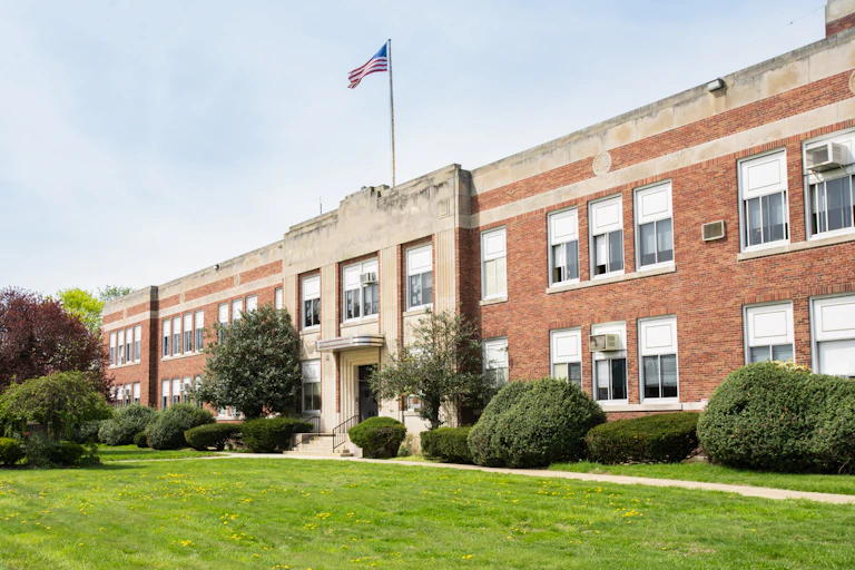 A traditional looking two-story school building with a rooftop flag blowing in the wind on a relatively sunny day.