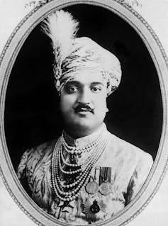 A black and white photo of a formal portrait of a man with a mustache and a headwrap with a feather.