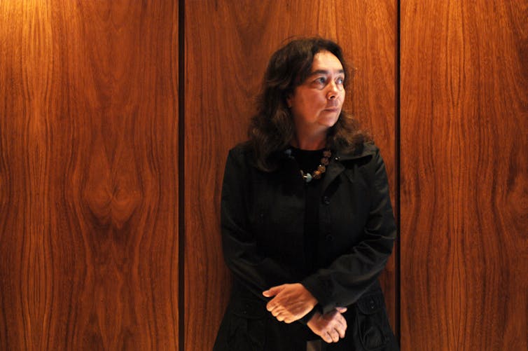 A woman with long hair, dressed in black, looks to her right, standing against a wood-grained wall