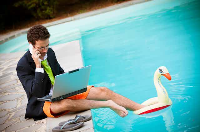 Man in suit jacket, tie, shirt & bathing suit on laptop by a pool