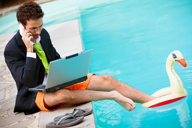 Man in suit jacket, tie, shirt & bathing suit on laptop by a pool