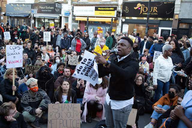 A crowd of protestors sit and stand as one man speaks with a poster in his hand. 
