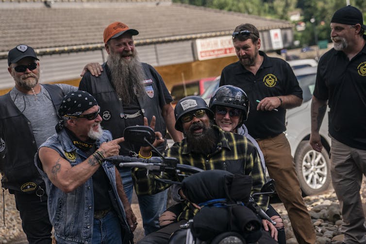 A group of men wear baseball hats, beards and casual clothing, including an official-looking Proud Boys shirt, as they smile and gather around two people on a motorcycle.