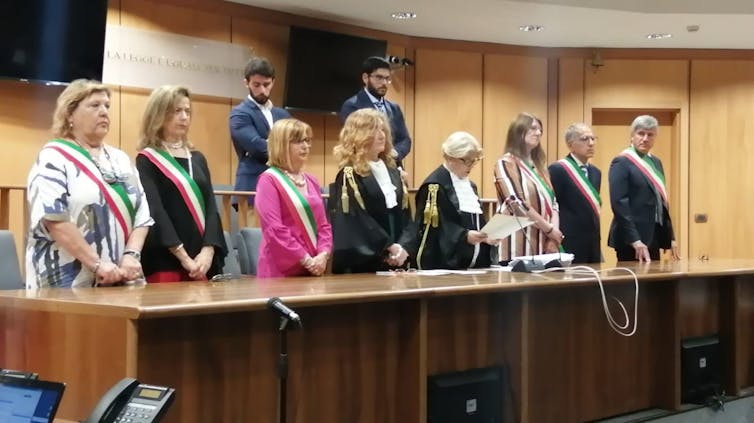 Jueces con cintas oficiales de pie detrás de un escritorio de madera en una sala de tribunal con paneles.