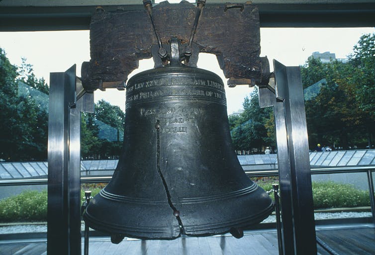 A large bell is displayed on a stand, with a shady courtyard in the background.